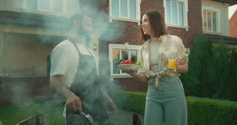 Happy brunette guy with a beard in a brown apron is engaged in barbecue while Stock Footage 278716306