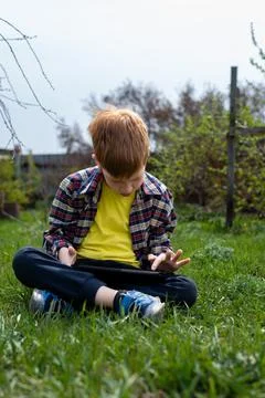 Happy child redhead boy playing on tablet or watching cartoons sitting on gre Stock Photos