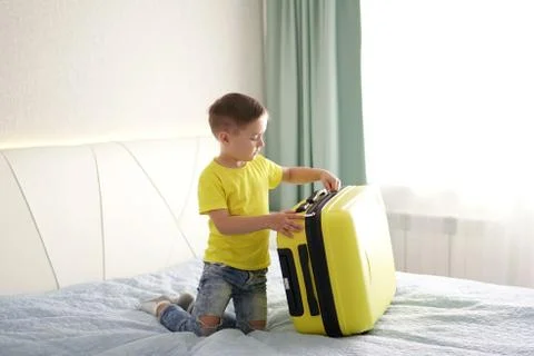 Happy child zips up a suitcase while sitting on the bed in the bedroom Stock Photos