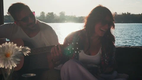 Happy couple laughing while playing guitar by the river Stock Footage 99978808