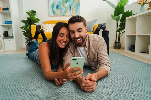 Happy couple lying on rug using smartphone together smiling at screen in cozy Stock Photos