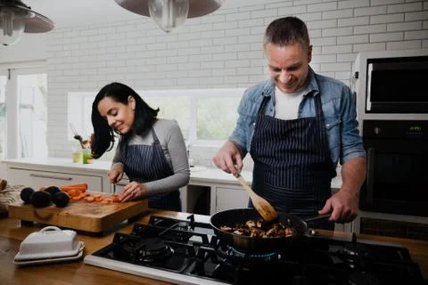 Happy couple preparing dinner while wife chops vegetables and husbands fries Stock Photos