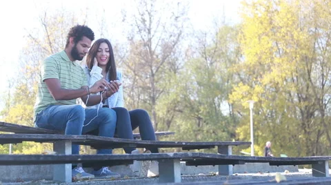 Happy couple sharing earphones while listening to music at park Stock Footage 59555223