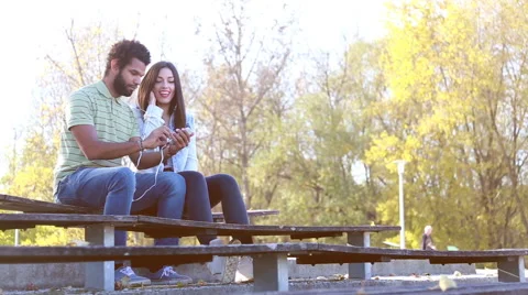 Happy couple sharing earphones while listening to music at park Stock Footage 59555234