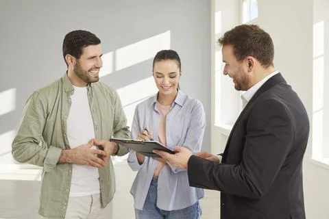 Happy couple signing contract with real estate agent in bright empty room after Stock Photos