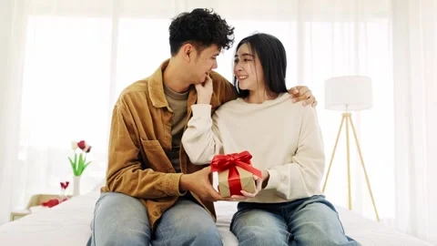 A happy couple sits on a bed, exchanging a gift wrapped with a red ribbon Видео 328076863