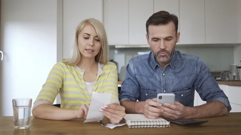 Happy couple sitting at dining table with phone and bills reviewing home budg Stock Footage 114607831