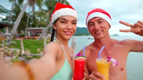 Happy couple smiling at camera taking self-portrait wearing santa hats on beach 스톡 동영상 119587053