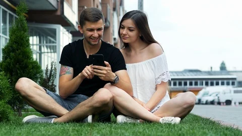 Happy couple using a mobile phone while sitting on the grass, online shopping Stock Footage 121678296