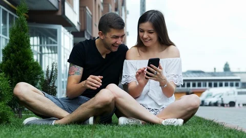 Happy couple using a mobile phone while sitting on the grass, online shopping Stock Footage 121678459
