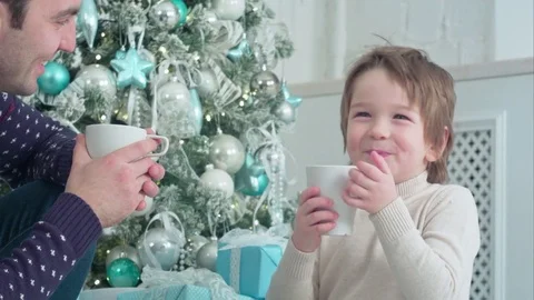 Happy dad and son with cups of tea talking near Christmas tree at home Vídeos de archivo 70404380