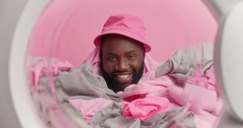 Happy dark skinned bearded guy wears pink panama on head poses through laundry Stock Footage 156633389