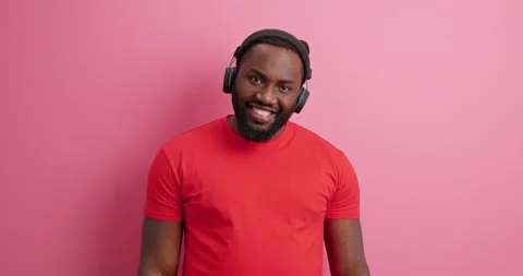 Happy dark skinned guy gestures with hand smiles carefree enjoys favorite audio Stock Footage 151616640