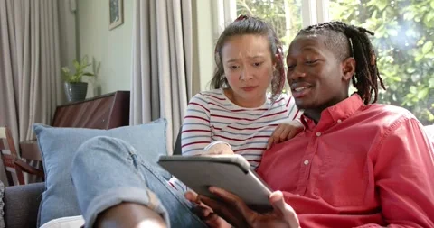 Happy diverse couple sitting on couch using tablet in sunny living room, slow Stock Footage 343238306