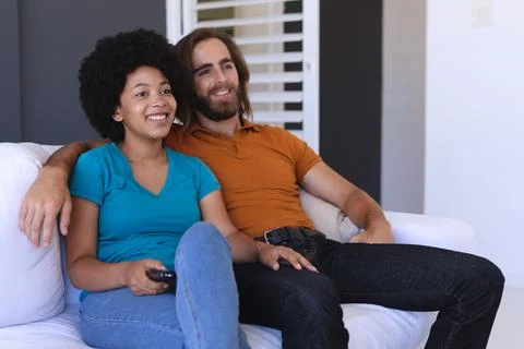Happy diverse couple sitting on sofa embracing and watching tv Stock Photos