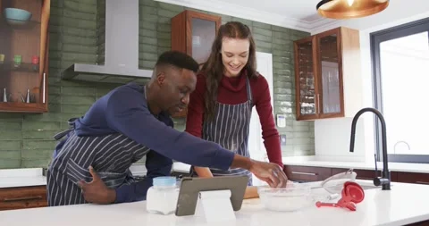 Happy diverse couple standing in kitchen, using tablet and preparing dough,slow Stock Footage 344338888