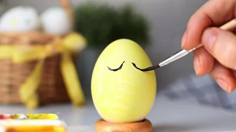 Happy Easter concept. Young womans hand draws a cute face on yellow egg. 스톡 동영상 234686698