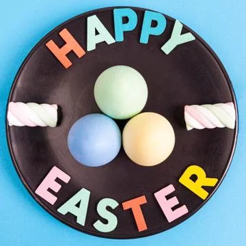 Happy Easter text with colorful eggs and marshmallows arranged on black plate Stock Photos