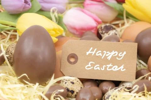 Happy Easter written on a label with chocolate eggs and pastel colored tulips Stock Photos