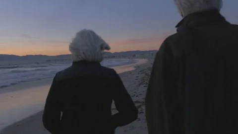 Happy elderly 60s couple walking and pointing along upscale beachfront at sunset Stock Footage 74434060