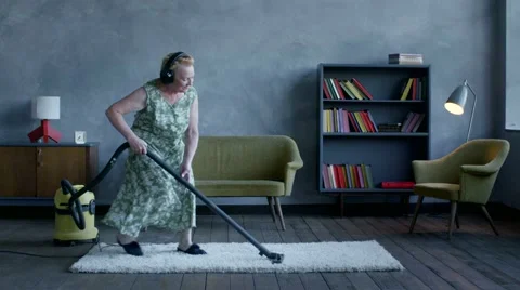 Happy elderly woman listening music on headphones and dancing with a vacuum Stock Footage