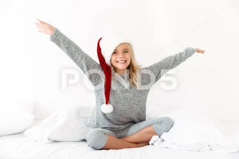Happy exited blonde girl in Santa's hat sitting on bed with raised arms, look Stock Photos
