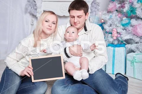 Happy family holding a blank Christmas card, sitting on the carpet in a cozy Stock Photos