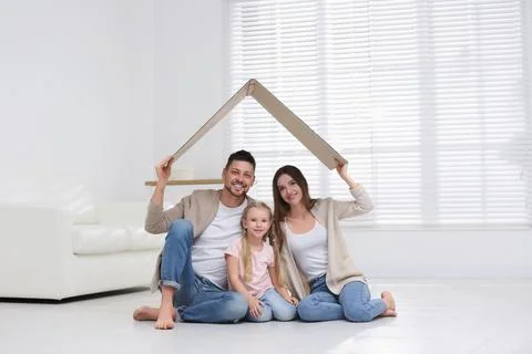 Happy family sitting under cardboard roof at home. Insurance concept Stock Photos