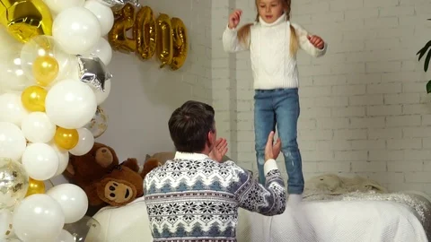Happy father in Christmas sweater catching adorable daughter jumping on bed in Stock Footage 98661663