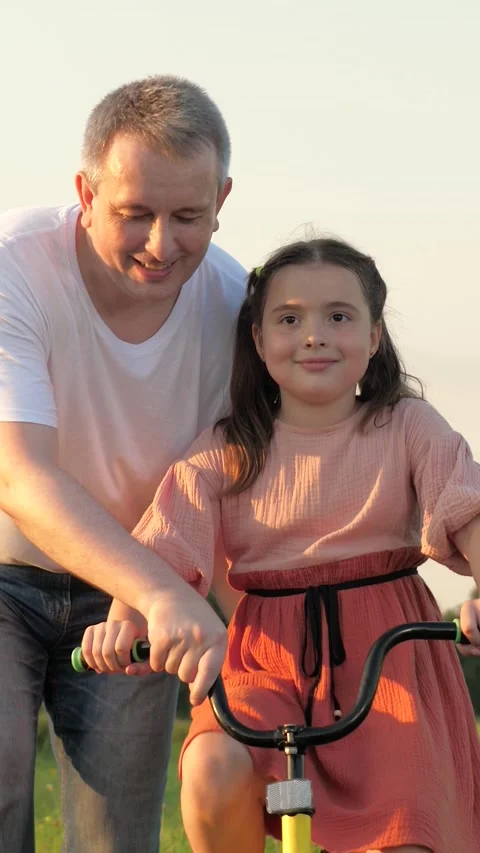 Happy father supports bicycle while little girl learning to ride in green field Stock Footage 274504125