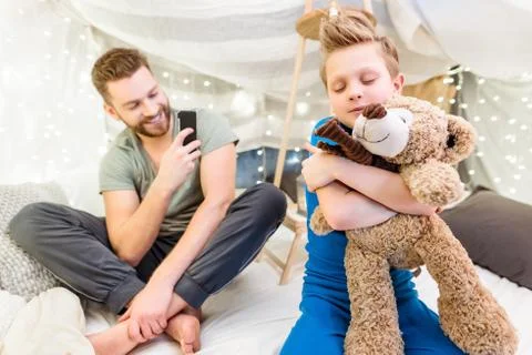 Happy father using smartphone while little son hugging with teddy bear Fotos Stock