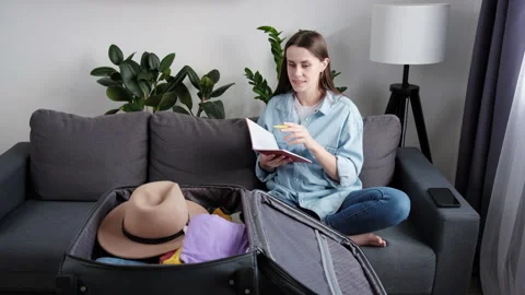 Happy female sit on couch take note and check list, prepare clothes into luggage Stock Footage 223360835