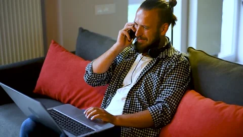 Happy freelancer talking on mobile phone using laptop on sofa in apartment. Video stock 136549720