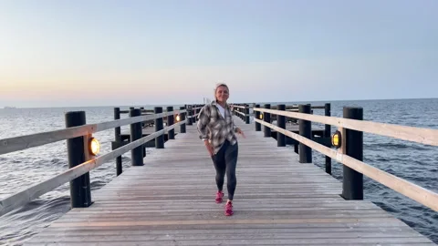 Happy, funny, dancing middle-aged woman wearing a shirt on a long wooden pier on Vídeo Stock 337736774