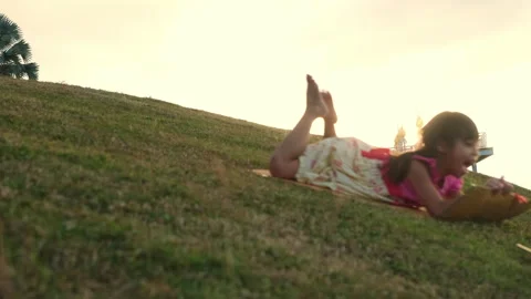 Happy girl slides down from grassy hill sitting on cardboard boxes at park Stock Footage