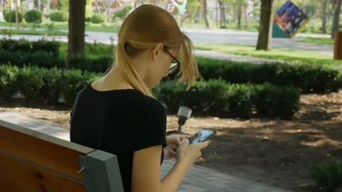 Happy girl student using a smart phone in a city park sitting on a bench, you Stockbeeldmateriaal 114178989