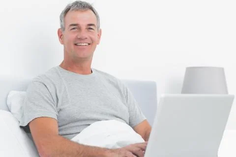 Happy grey haired man using his laptop in bed Stock Photos