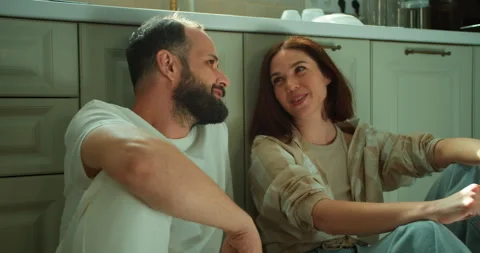 Happy guy with a beard in a white T-shirt sits on the floor in the kitchen with Stock Footage 280251005