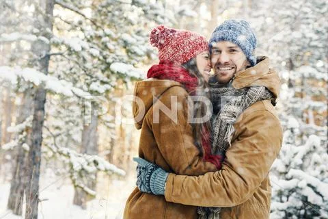 Happy guy looking at camera while embracing his girlfriend in snowfall Stock Photos