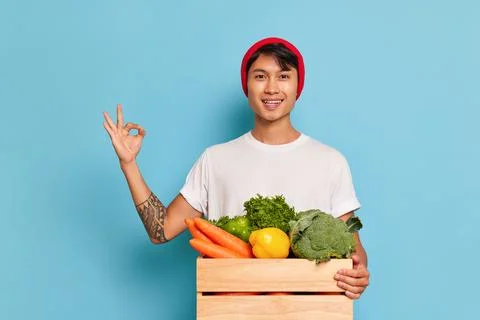 Happy guy in red hat and white t-shirt poses against blue background, box with Stock-Fotos