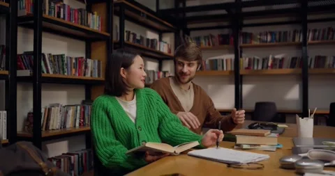 Happy guy talking to girl while studying in front of books at table in library Stock Footage 313347729