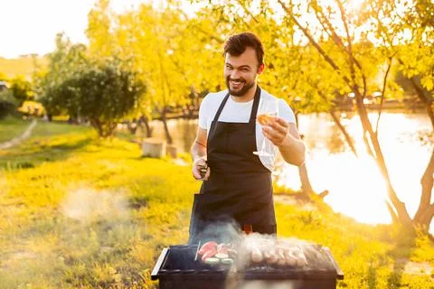 Happy guy toasting to camera while grilling sausages and vegetables in park Stock Photos