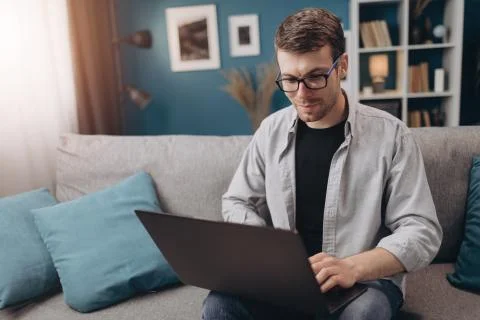 Happy guy using laptop while sitting on grey couch Stock Photos