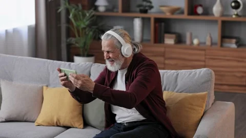 Happy handsome elderly gray-haired man in white headphones holding smartphone Stock Footage 198429771