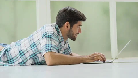 Happy handsome man lying down on floor using laptop computer at home Stock Footage 134399167