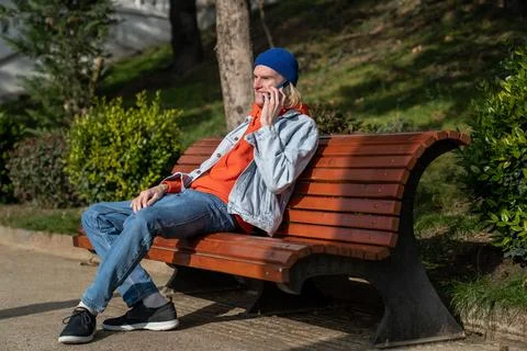 Happy hipster guy talking on mobile phone while relaxing on bench in park on Stock Photos