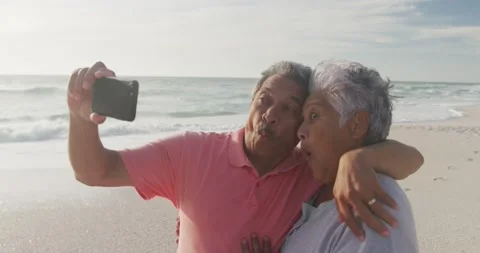 Happy hispanic senior couple making funny faces, taking selfie on beach at Stock Footage 335198155