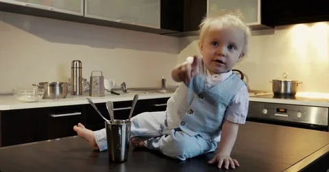 A happy little chef chef sits on a table and plays with metal spoons, mugs. Stock Footage 84629117