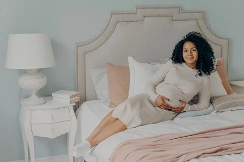 Happy lovely pregnant female relaxing on bed at home and touching her belly Stock Photos