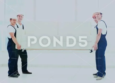 Happy male movers in uniform carrying white sofa Stock Photos